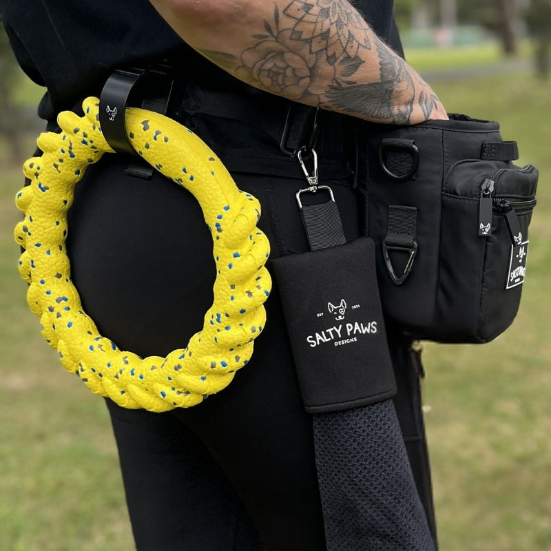 A close-up lifestyle shot of a person wearing a black Salty Paws Designs SnapLock Pro Training Pouch. A bright yellow textured dog ring is attached to the belt, and a black pouch with the white 'SALTY PAWS DOG' logo is attached.