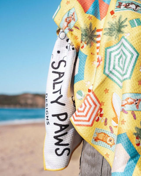 Salty Paws Designs Life's a Beach dog towel in yellow draped over a post at the beach, showing the vibrant beach umbrella print and Salty Paws Designs branding