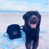 Chocolate Labrador sitting on the Salty Paws Designs Pool Party adventure dog towel in blue at the beach, with a Salty Paws Designs bag beside it